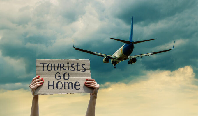 Woman greets a plane with cardboard sign "Tourists go home", stop overtourism concept; photo by sergii.kl.ua, Depositphotos.