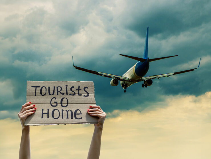 Woman greets a plane with cardboard sign "Tourists go home", stop overtourism concept; photo by sergii.kl.ua, Depositphotos.