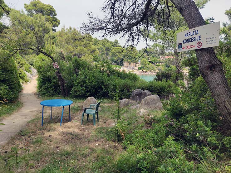 Blue round table and green plastic chair at one of the two check-points at FKK Kandarola Beach on Rab Island, Croatia, a naturist beach that's been managed under concession since 2015 by Josip Jurešić, Boja d.o.o., who charges 4 euros for passage, as indicated on the sign nailed to the tree; photo by Ivan Kralj