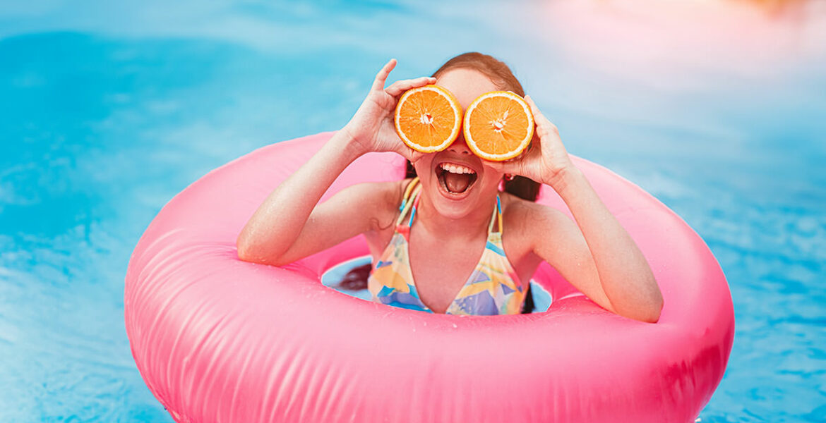 Cheerful girl in swimsuit covering eyes with sliced oranges while having fun in pool with pink inflatable ring; it's time to get essential products for the best season ever, with these summer must-haves! Photo by Kegfire, Depositphotos.
