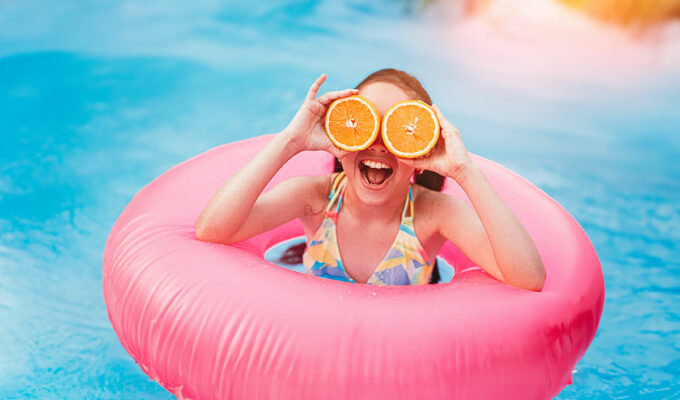 Cheerful girl in swimsuit covering eyes with sliced oranges while having fun in pool with pink inflatable ring; it's time to get essential products for the best season ever, with these summer must-haves! Photo by Kegfire, Depositphotos.