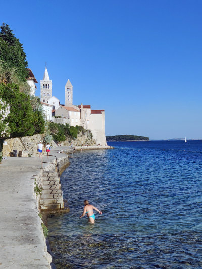 Swimmer exiting the sea beneath the walls of Rab town in Croatia, with church bell towers in the background; photo by Ivan Kralj.