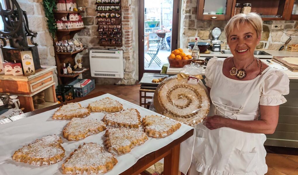Baker Ružica Ribarić holding a famous traditional spiral Rab cake, standing next to a display of heart-shaped cakes, at Kuća rabske torte in Rab, the central institution promoting rapska torta, Croatian almond delicacy that was first baked for Pope Alexander III; photo by Ivan Kralj.