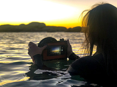 Woman in the sea taking a photograph of the sunset with her mobile phone in an underwater housing by Gdome.