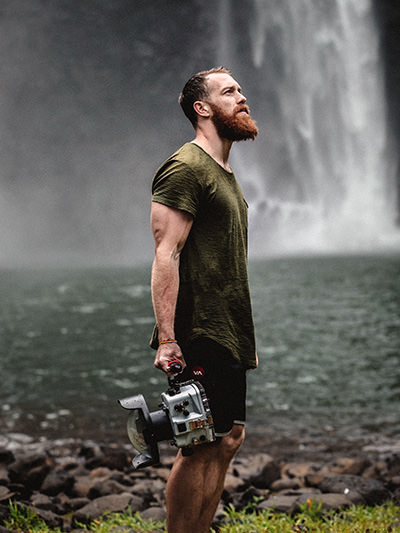 Muscular bearded man in front of a waterfall, holding a camera in a bulky underwater housing in his hand; photo by Jakob Owens, Unsplash.