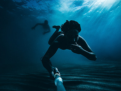 Two divers under water with one taking a photograph with a camera on a selfie stick, the image is in blue tone; photo by Daoud Abismail, Unsplash.
