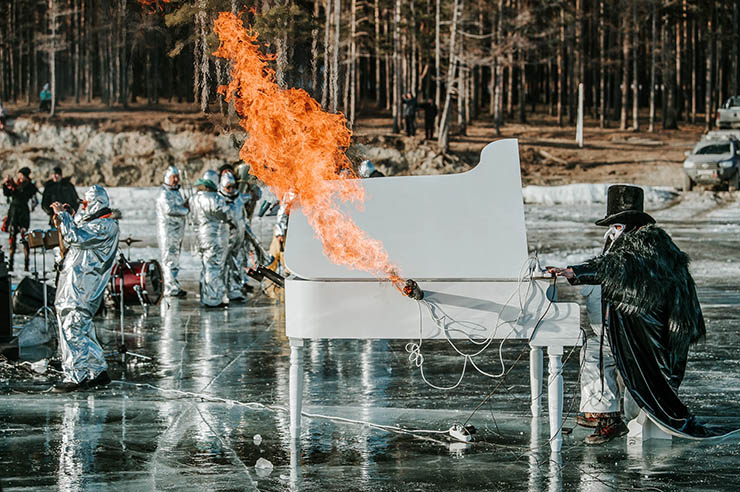 Man in mask and black cylinder hat playing a piano on the frozen Lake Baikal with fire blowing on the side of the instrument, part of the MadWay Rally, Mad Max-inspired event.