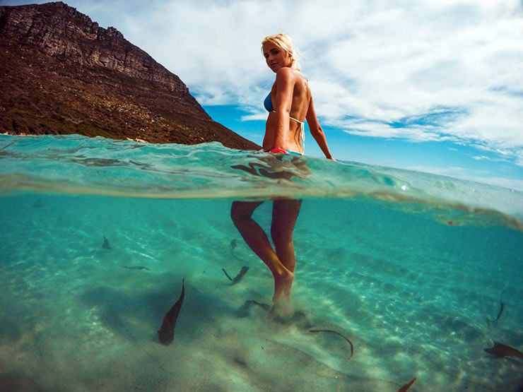 A woman in a bathing suit walking through the water with fish swimming below the surface, shot with split-level photography; photo by GDome.