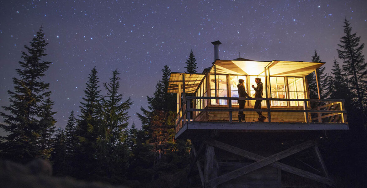 Women standing in illuminated cottage at starry night, a promo visual for Booking.com's Early 2024 Deals that enable discounts of minimum 15%.