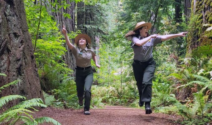 Two female rangers of National Park Service comically pointing at the forest while running through it; NPS humor safety marketing on Twitter.