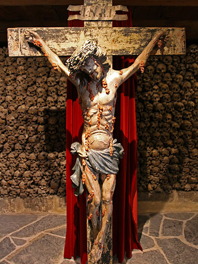 Crucifix of heavily-bleeding Jesus in the center of Beinhaus Leuk, with the wall of skulls and bones in the background, Walis, Switzerland; photo by Ivan Kralj.