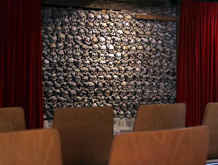 Chairs facing the wall full of skulls, framed by two red curtains, resembling a theater, photo taken in Beinhaus Leuk, Swiss ossuary in Wallis, by Ivan Kralj.