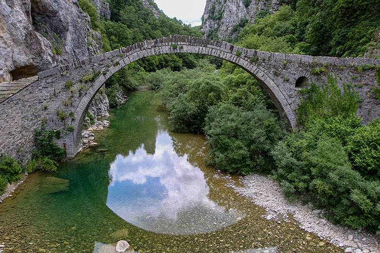 Kokkorou Bridge, a one-arch stone bridge in the mountainous landscape of Zagori, Epirus, Greece, reflecting in the river beneath it, and surrounded by lush greenery; photo by Ivan Kralj.