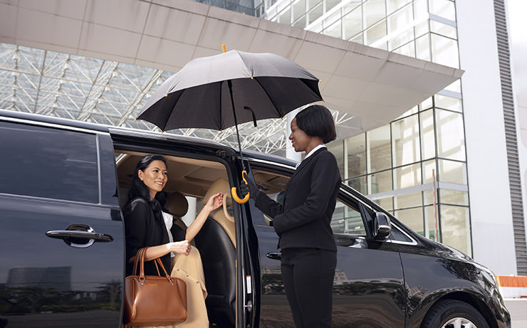 Businesswoman exiting a taxi cab at the airport while a member of staff is holding an umbrella/parasol for her convenience; image by Freepik.