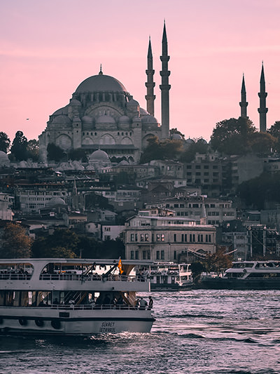 A mosque in Istanbul, Turkey (one of the best places to visit in 2024), as seen from water, with sunset-colored sky; photo by Ibrahim Guetar, Unsplash.