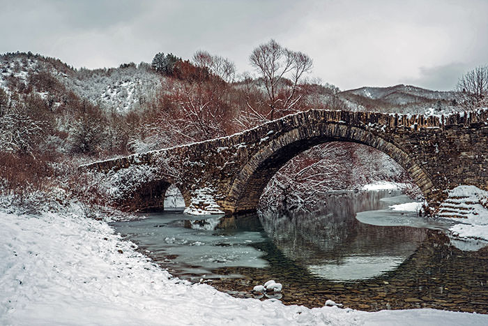 Milos Bridge in Kipoi, a village in Zagori, Greece, during winter, surrounded by snow-covered landscape; photo by Bob Jansen, Unsplash.