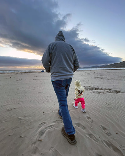 Tall dad Eric Castillo and tiny daughter Journey walking over the beach in hoodies.
