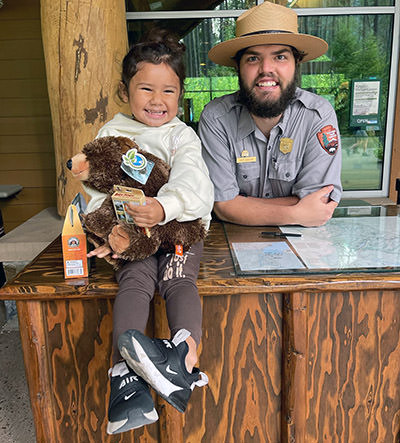 Journey Castillo (3) with a plush teddy bear, posing next to a park ranger in Glacier National Park, one of 63 U.S. national parks the toddler visited before turning 3 years old. 