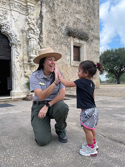 Three-year-old hiker Journey Castillo high-fiving a park ranger at San Antonio Missions National Park, one of the 63 national parks she visited in the United States.