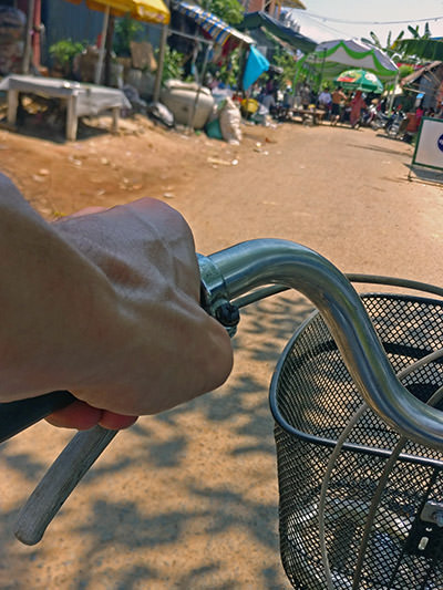 Hands on the handlebar of a bicycle, riding on a dusty street of Kampong Chhnang, Cambodia, with a celebration tent in the middle of the road; photo by Ivan Kralj.