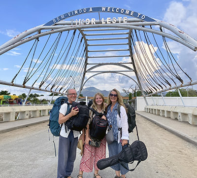 Theo Simon, Rosa, and Shannon Coggins standing with backpacks and a guitar at the entrance to East Timor, the country where their no-fly trip to Australia will come to an unsurmountable obstacle - the slow-travel family will be forced to take a plane.