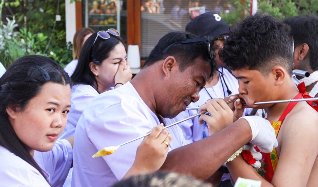 A man in gloves adjusting the spike piercing a cheek of a boy, while a woman is passing by with a reaction of shock, during Jay or Phuket Vegetarian Festival, also known as Thailand's self-mutilation festival; photo by Ivan Kralj.