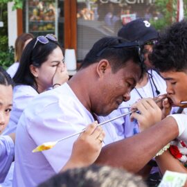 A man in gloves adjusting the spike piercing a cheek of a boy, while a woman is passing by with a reaction of shock, during Jay or Phuket Vegetarian Festival, also known as Thailand's self-mutilation festival; photo by Ivan Kralj.