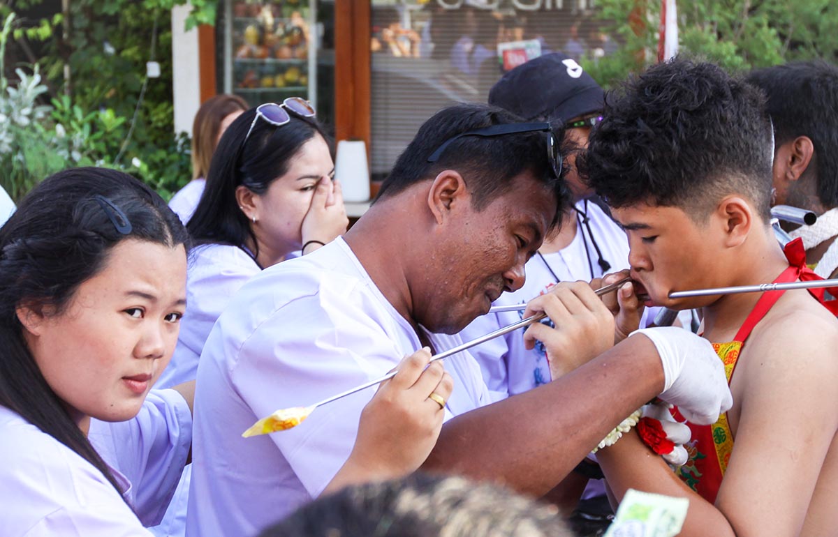 A man in gloves adjusting the spike piercing a cheek of a boy, while a woman is passing by with a reaction of shock, during Jay or Phuket Vegetarian Festival, also known as Thailand's self-mutilation festival; photo by Ivan Kralj.