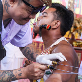 A piercer piercing the arm skin of a man, a ma song spirit medium at Jor Soo Goong Naka Shrine, during Jay or Phuket Vegetarian Festival, also known as Thailand's self-mutilation festival; photo by Ivan Kralj.