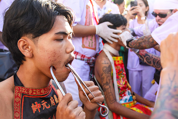A young man holding hooks piercing his cheeks, while another man is getting pierced in the background, at Jor Soo Goong Naka Shrine, during Jay or Phuket Vegetarian Festival, also known as Thailand's self-mutilation festival; photo by Ivan Kralj.