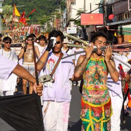 A man with swords piercing his cheeks, walking in a street procession through Phuket during Jay or Vegetarian Festival, also known as Thailand's self-mutilation festival, with deity sedan chair in the background; photo by Ivan Kralj.