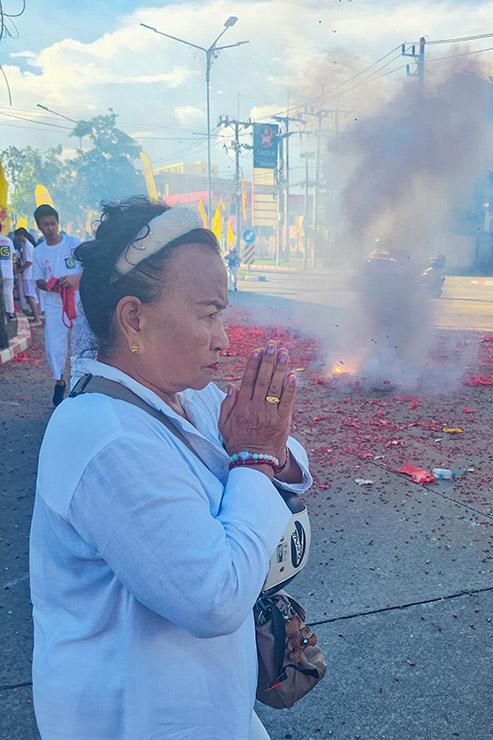 A woman standing in the street, with hands in a praying position, while firecrackers explode and create a smoke in the background, during Jay or Phuket Vegetarian Festival; photo by Ivan Kralj.