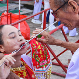 An older man adjusting a metal rod piercing a cheek of a woman, a ma song spirit medium at Jor Soo Goong Naka Shrine, during Jay or Phuket Vegetarian Festival, also known as Thailand's self-mutilation festival; photo by Ivan Kralj.