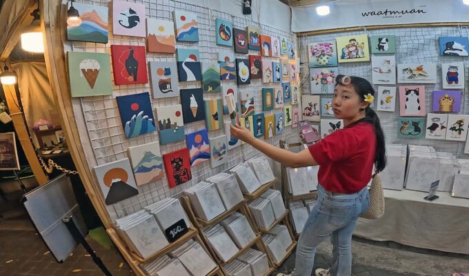 Young woman with a curler in her hair observing a painting being sold at the White Market, one of Nimman night markets in Chiang Mai, Thailand; photo by Ivan Kralj.