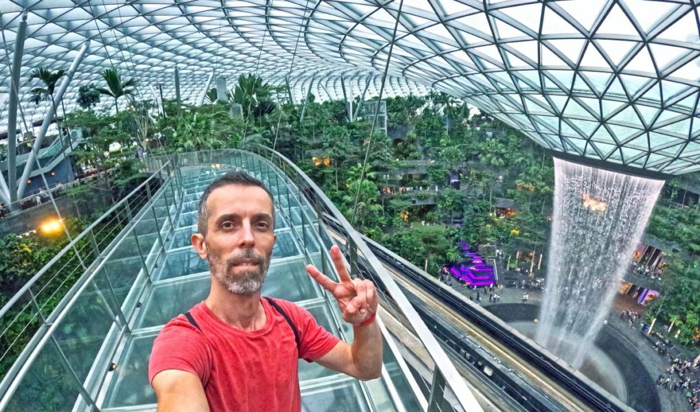 Travel blogger Ivan Kralj standing on Mastercard Canopy Bridge with Rain Vortex in the background, the world's largest indoor waterfall in a stunning Jewel complex of Singapore Changi Airport; photo by Ivan Kralj.
