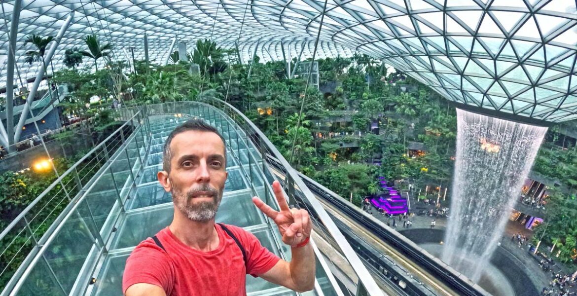Travel blogger Ivan Kralj standing on Mastercard Canopy Bridge with Rain Vortex in the background, the world's largest indoor waterfall in a stunning Jewel complex of Singapore Changi Airport; photo by Ivan Kralj.