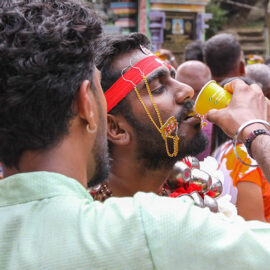 A man assisting Hindu devotee with drinking, as he has alavu kavadi (pierced cheeks and tongue), during Thaipusam in Penang, Malaysia; photo by Ivan Kralj.