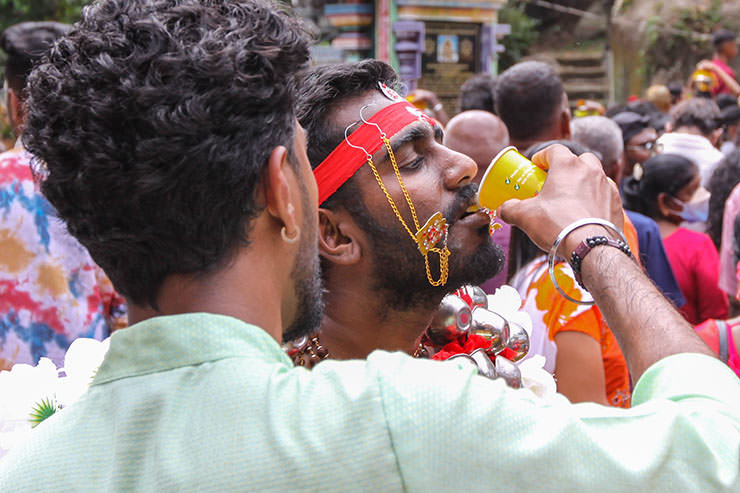 A man assisting Hindu devotee with drinking, as he has alavu kavadi (pierced cheeks and tongue), during Thaipusam in Penang, Malaysia; photo by Ivan Kralj.