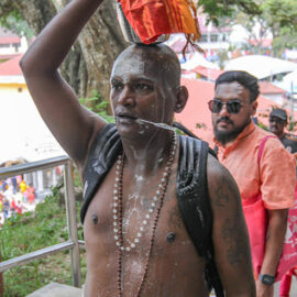 A Hindu man with pierced cheeks carrying a milk pot (paal kudam) on his head, while milk pours down his bare torso, during Thaipusam Festival in Penang, Malaysia; photo by Ivan Kralj.