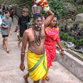 A shirtless Hindu man with pierced cheeks and tongue (alavu kavadi) carrying a milk pot (paal kudam) on his head, during Thaipusam Festival in Penang, Malaysia; photo by Ivan Kralj.