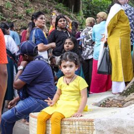 A young girl dressed in all yellow, sitting on the floor holding a V-sign as she poses for the camera, while the adult crowd stands in the background, stuck on the staircase towards the Waterfall Temple in Penang, Malaysia, during Thaipusam; photo by Ivan Kralj.