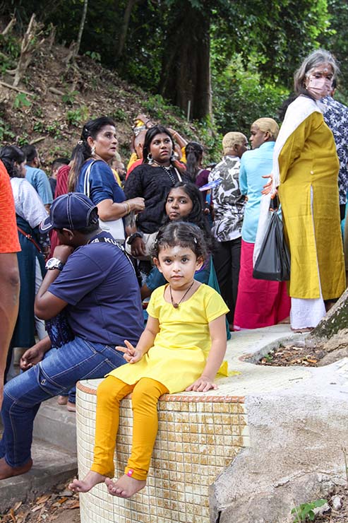 A young girl dressed in all yellow, sitting on the floor holding a V-sign as she poses for the camera, while the adult crowd stands in the background, stuck on the staircase towards the Waterfall Temple in Penang, Malaysia, during Thaipusam; photo by Ivan Kralj.