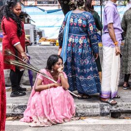 A young woman sitting on the sidewalk while holding peacock feathers, in a reflective pose, during Thaipusam in Penang, Malaysia; photo by Ivan Kralj.