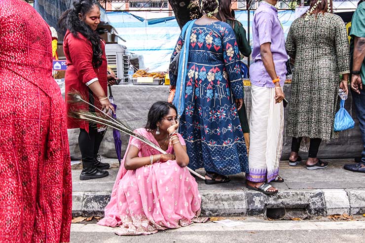 A young woman sitting on the sidewalk while holding peacock feathers, in a reflective pose, during Thaipusam in Penang, Malaysia; photo by Ivan Kralj.