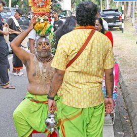 A seated Hindu devotee holding paal kudam milk pot on his head, with alavu kavadi - pierced cheeks and forehead, during Thaipusam in Penang, Malaysia; photo by Ivan Kralj.
