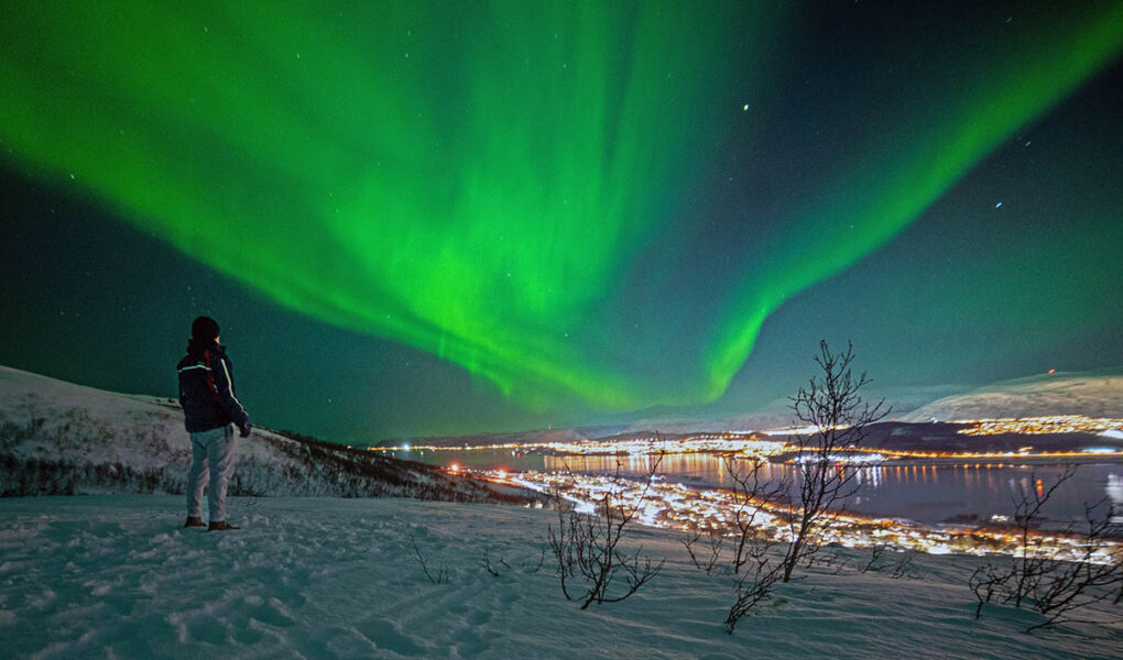 Man standing in nature at night with Tromso, Norway, city lights in the distance, and green northern lights in the sky; photo by Sven Pieren / Unsplash.