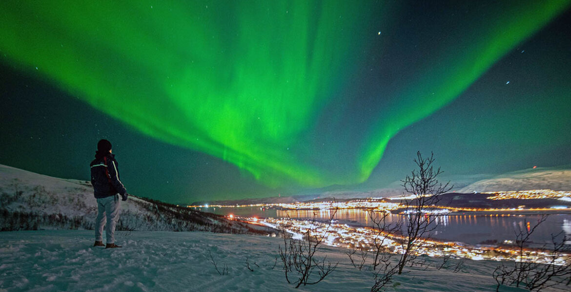 Man standing in nature at night with Tromso, Norway, city lights in the distance, and green northern lights in the sky; photo by Sven Pieren / Unsplash.