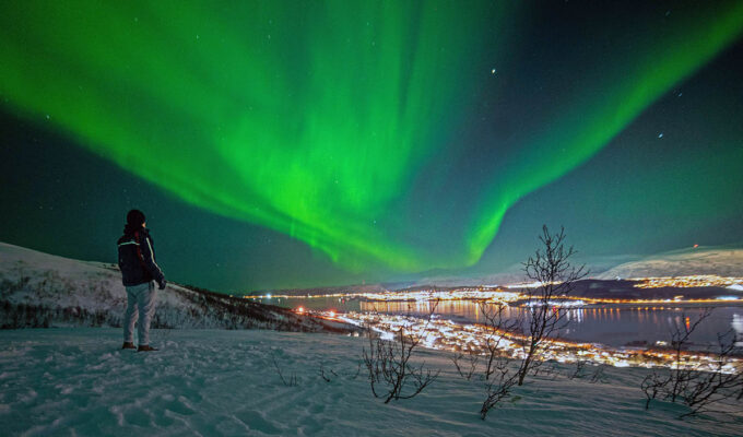 Man standing in nature at night with Tromso, Norway, city lights in the distance, and green northern lights in the sky; photo by Sven Pieren / Unsplash.