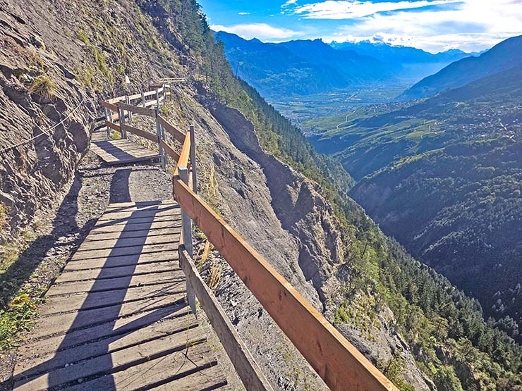 Wooden pathway zigzagging along the cliff of Prabé Mountain, following the Bisse du Torrent Neuf hike route, Saviese, Switzerland, with Alps in the background; photo by Ivan Kralj.