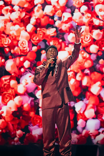 Congolese-Dutch musician Claude performing his song "C'est la vie" on Eurovision in Basel, Switzerland, against a backdrop of pink roses; photo by Sarah Louise Bennett / EBU.